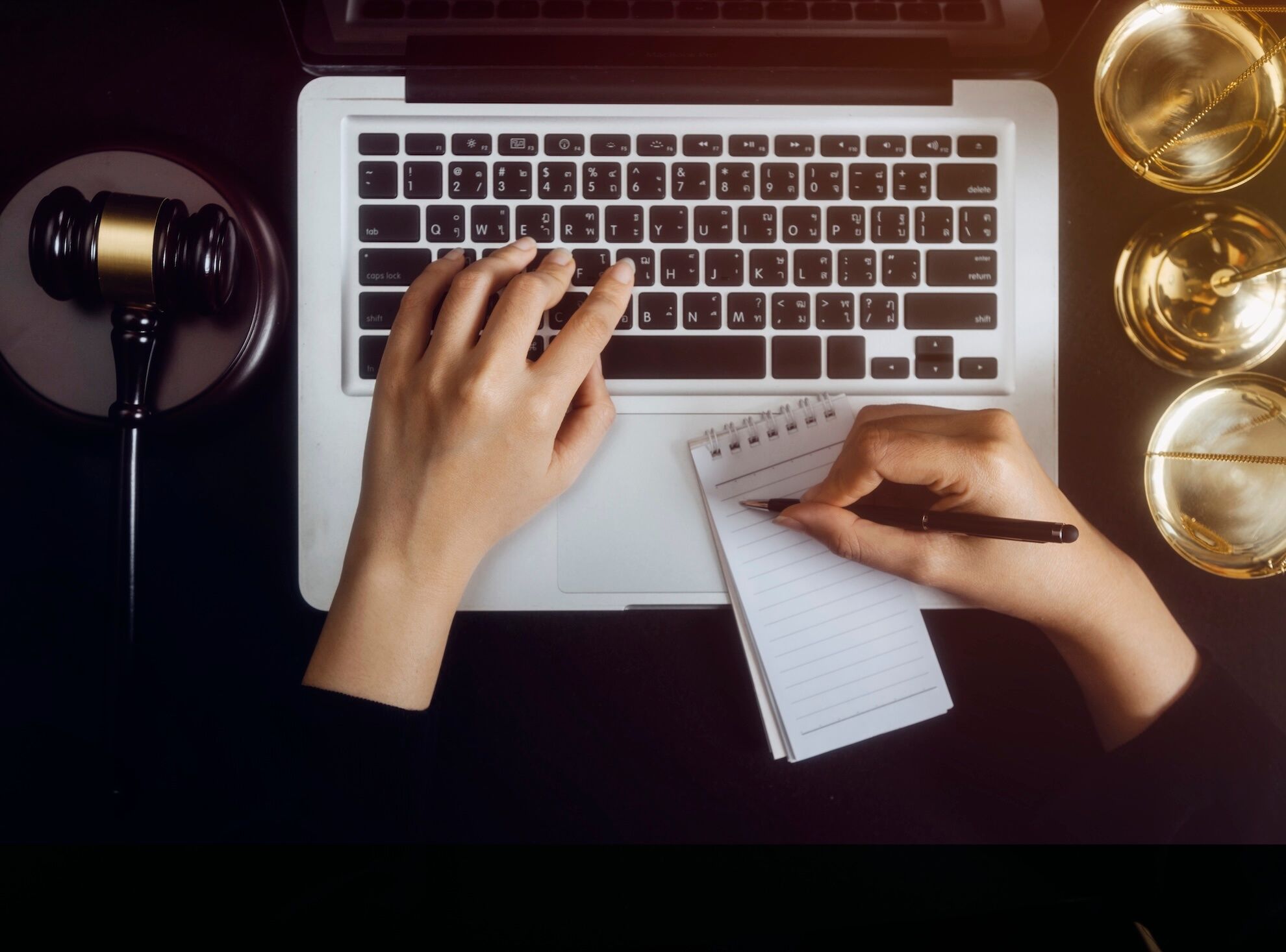 Una persona escribe en una computadora portátil mientras escribe en un bloc de notas, con un mazo y gafas cerca del escritorio.