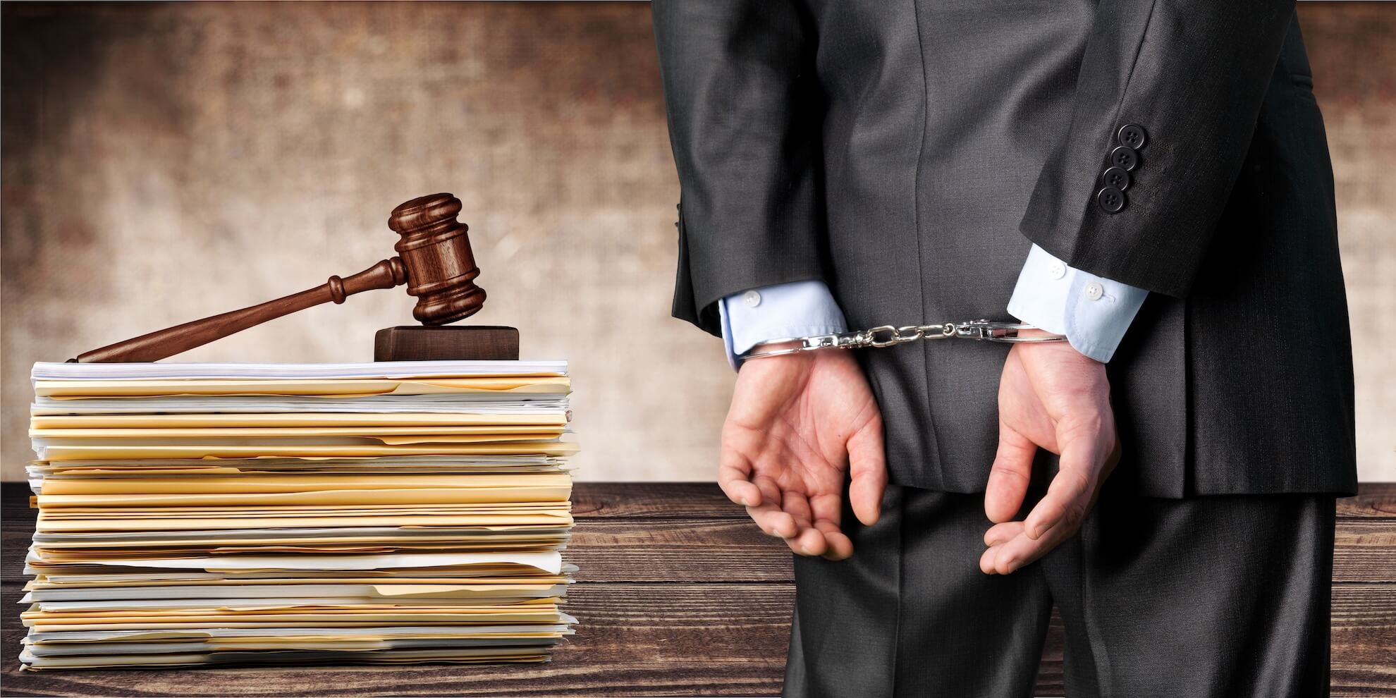 Un hombre de traje está esposado, de pie junto a un mazo y una pila de documentos legales sobre un escritorio. Derecho Penal. Elena Giménez Abogada
