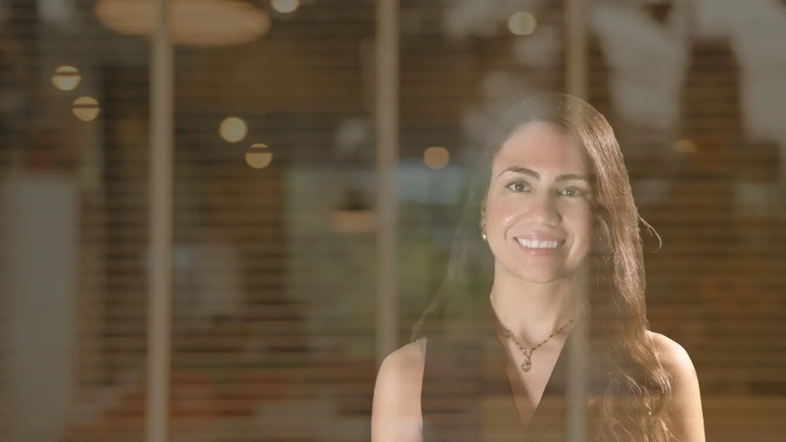 Una mujer de pelo largo sonríe detrás de una ventana. En el cristal se ven reflejos de luces y un interior borroso. Elena Giménez Abogada
